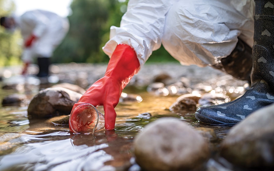Scientists in personal protective equipment collect water samples in cups from a stream