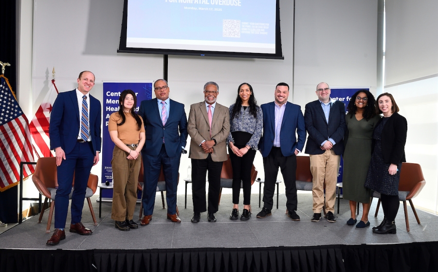 Nine adults standing on stage, including four panelists and a moderator. They're all in business attire.