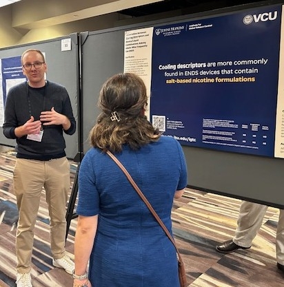 A man in a blue sweater and tan pants speaks to a woman in a blue top about his research