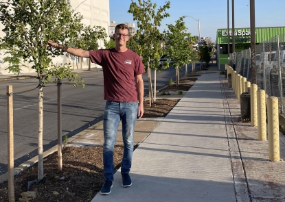 Man in red shirt and jeans standing on a sidewalk with his hand on a tree