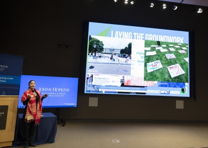 Dark haired woman wearing red scarf and black shirt presenting in front of screen in a lecture hall.