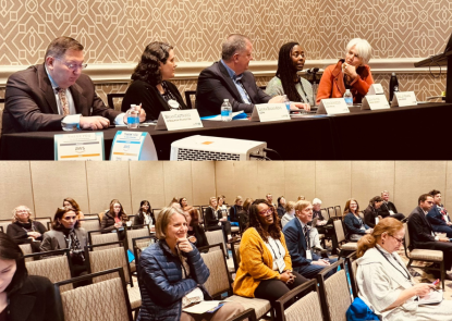 5 adults sitting at a table for a panel discussion. Room of people sitting in chairs.