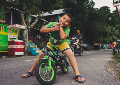 kid with a green shirt sitting on a green bike while leaning forward to look into the camera