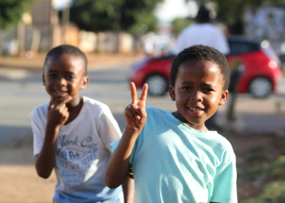 Two young boys outside, smiling, one is holding up two fingers in a "peace" sign