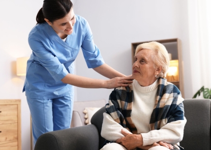 A woman wearing a blue nurse uniform places a hand on an elderly woman who is seated on a couch. They are both grinning.